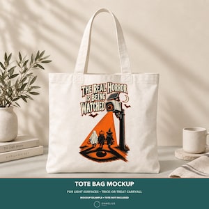 May include: A white canvas tote bag featuring a graphic print of a security camera shining an orange light on three figures dressed as a ghost, a pumpkin, and a witch. The text reads, THE REAL HORROR IS BEING WATCHED, with bats and a crescent moon.
