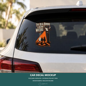 May include: A car decal featuring a security camera casting an orange light beam onto three figures dressed as a ghost, a pumpkin, and a witch. The text reads THE REAL HORROR IS BEING WATCHED, with a large eye graphic at the bottom of the design.
