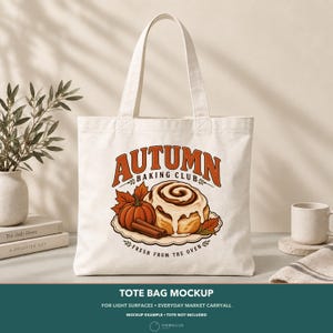 May include: A cream-colored canvas tote bag featuring a graphic print of a cinnamon roll, a pumpkin, and cinnamon sticks. The text reads "AUTUMN BAKING CLUB" in large orange letters and "FRESH FROM THE OVEN" in smaller black text below the illustration.