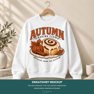 May include: A white crewneck sweatshirt featuring a graphic print of a cinnamon roll, a pumpkin, and cinnamon sticks. The text reads AUTUMN BAKING CLUB and FRESH FROM THE OVEN. The design includes orange and brown autumn leaves on a scalloped plate.