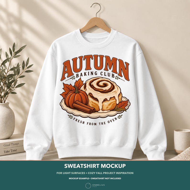May include: A white crewneck sweatshirt featuring a graphic print of a cinnamon roll, a pumpkin, and cinnamon sticks. The text reads AUTUMN BAKING CLUB and FRESH FROM THE OVEN. The design includes orange and brown autumn leaves on a scalloped plate.