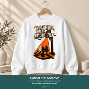 May include: A white crewneck sweatshirt featuring a graphic print of a security camera with an orange beam of light shining on three children in Halloween costumes. The text reads, THE REAL HORROR IS BEING WATCHED, with bats and a crescent moon.