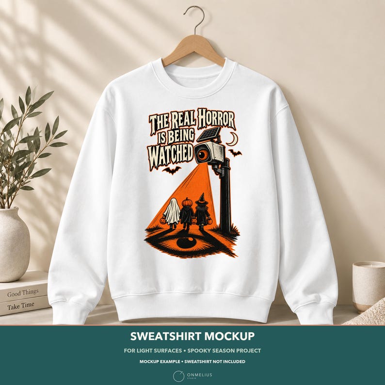 May include: A white crewneck sweatshirt featuring a graphic print of a security camera with an orange beam of light shining on three children in Halloween costumes. The text reads, THE REAL HORROR IS BEING WATCHED, with bats and a crescent moon.