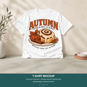 May include: A white short-sleeved t-shirt featuring a graphic of a cinnamon roll, a pumpkin, and cinnamon sticks. The text reads AUTUMN BAKING CLUB and FRESH FROM THE OVEN. The design includes orange and brown autumn leaves on a scalloped edge base.