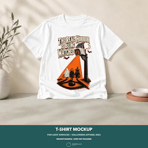 May include: A white t-shirt featuring a graphic print of a security camera casting an orange light beam onto three figures in Halloween costumes. The text reads, THE REAL HORROR IS BEING WATCHED, accompanied by a crescent moon and bats.