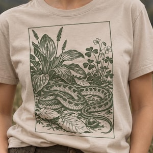 Puede incluir: Una camiseta beige de manga corta con un estampado gráfico rectangular en tinta verde oscuro. La ilustración muestra una serpiente enroscada rodeada de elementos botánicos detallados, incluyendo hojas anchas, tallos altos y flores pequeñas.