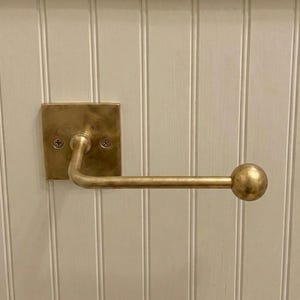 May include: A brass toilet paper holder featuring a square wall-mounted backplate with two visible screws and a horizontal arm that extends outward, ending in a spherical finial. The metal has a brushed, matte finish with warm golden tones.