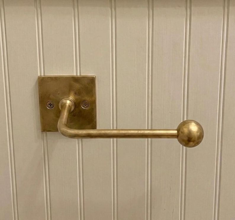 May include: A brass toilet paper holder featuring a square wall-mounted backplate with two visible screws and a horizontal arm that extends outward, ending in a spherical finial. The metal has a brushed, matte finish with warm golden tones.