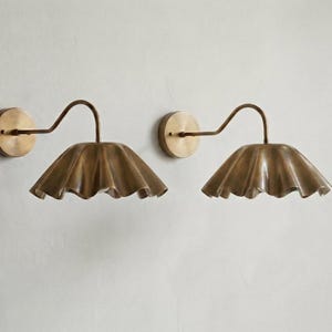 May include: A pair of wall-mounted lamps featuring circular brass backplates, curved arms, and wide, ruffled, bell-shaped shades with a metallic brass finish. The design includes a smooth, polished texture on the hardware and a fluted edge.
