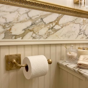 May include: A brass toilet paper holder mounted on a white beadboard wall, holding a roll of white toilet paper. Nearby on a marble countertop sits a clear glass jar with a lid, a white candle, and a folded beige cloth with a letter B.