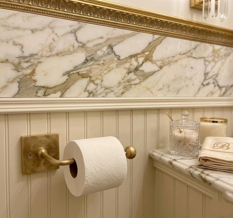 May include: A brass toilet paper holder mounted on a white beadboard wall, holding a roll of white toilet paper. Nearby on a marble countertop sits a clear glass jar with a lid, a white candle, and a folded beige cloth with a letter B.