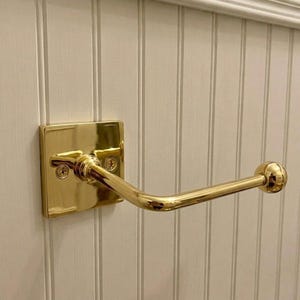 May include: A polished brass toilet paper holder featuring a square wall-mounted backplate with two visible screws and a horizontal arm that extends outward, ending in a rounded knob to secure a roll of paper in place.