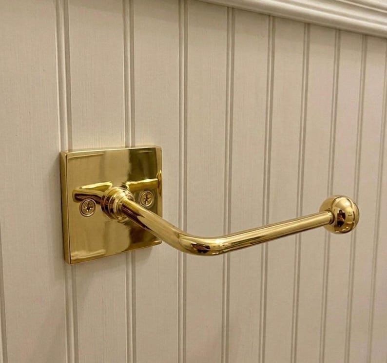 May include: A polished brass toilet paper holder featuring a square wall-mounted backplate with two visible screws and a horizontal arm that extends outward, ending in a rounded knob to secure a roll of paper in place.