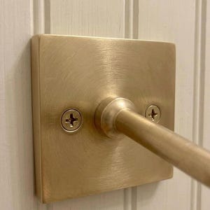 May include: A square, brushed brass wall plate with a central cylindrical rod extending outward. The plate features a circular mounting base and is secured to the surface by two visible Phillips head screws, one on each side of the central rod.