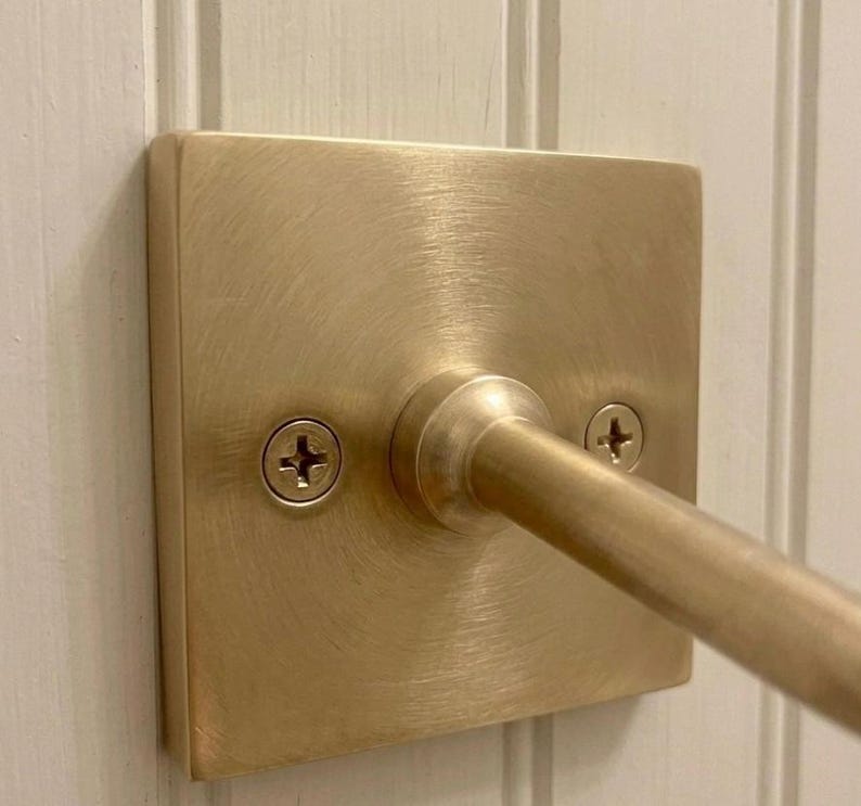 May include: A square, brushed brass wall plate with a central cylindrical rod extending outward. The plate features a circular mounting base and is secured to the surface by two visible Phillips head screws, one on each side of the central rod.
