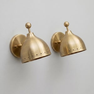 May include: Two brass wall sconces with a classic design. Each sconce features a round base, a conical shade with small perforations, and a small ball finial. The sconces are mounted on a light gray wall.