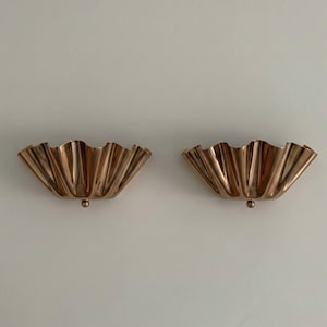 May include: Two copper-coloured wall sconces with a scalloped, fan-like design. Each sconce has a small, round base. The sconces are mounted on a white wall. The design is reminiscent of Art Deco style.