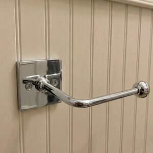 May include: A polished chrome toilet paper holder featuring a square wall-mounted base plate with two visible screws and a single horizontal arm that extends outward, ending in a rounded spherical finial for securing a roll of paper.