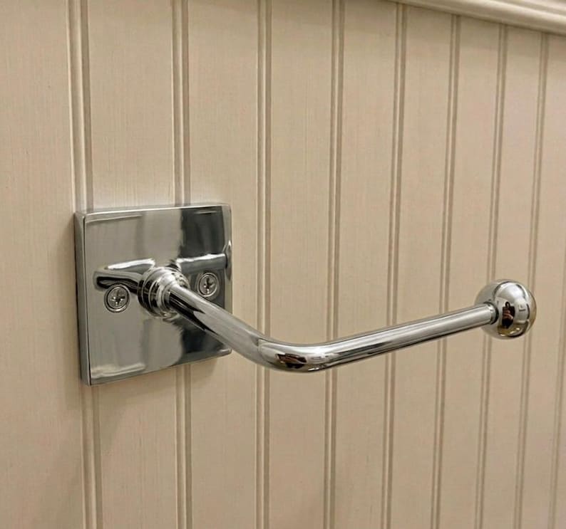 May include: A polished chrome toilet paper holder featuring a square wall-mounted base plate with two visible screws and a single horizontal arm that extends outward, ending in a rounded spherical finial for securing a roll of paper.