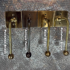 May include: Four metal wall hooks with square backplates and curved arms ending in spherical knobs. The finishes are labeled as Polished Brass, Antique Patina, Raw Brass, and Polished Brass, showing variations in metallic color and surface texture.