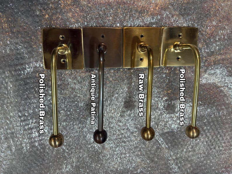 May include: Four metal wall hooks with square backplates and curved arms ending in spherical knobs. The finishes are labeled as Polished Brass, Antique Patina, Raw Brass, and Polished Brass, showing variations in metallic color and surface texture.