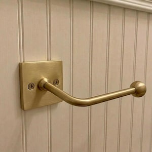 May include: A brushed brass wall-mounted hook featuring a square backplate with two visible screws and a horizontal arm that extends outward, ending in a rounded ball finial. The hardware has a metallic finish and a simple, functional design.