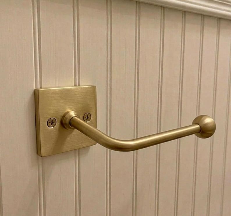 May include: A brushed brass wall-mounted hook featuring a square backplate with two visible screws and a horizontal arm that extends outward, ending in a rounded ball finial. The hardware has a metallic finish and a simple, functional design.