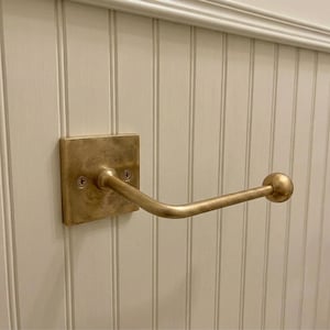 Könnte beinhalten: Ein an der Wand montierter Toilettenpapierhalter aus Messing mit einer quadratischen Rückplatte, zwei sichtbaren Schrauben und einem horizontalen Arm, der nach außen ragt und in einem runden Kugelabschluss endet. Das Metall hat ein mattes Finish.
