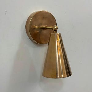 May include: A wall-mounted light fixture featuring a circular brass backplate and a conical brass shade. The shade is connected to the backplate by a horizontal arm with a swivel joint, allowing for adjustable positioning of the light direction.
