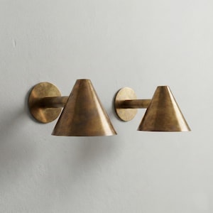 May include: Two brass wall sconces. Each sconce features a circular base, a horizontal arm, and a conical shade. The sconces are made of a warm, metallic brass material and are mounted on a light-colored wall.