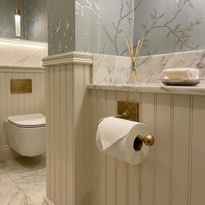 May include: A bathroom interior featuring a wall-mounted white toilet with a brass flush plate, a white toilet paper roll on a brass holder, and a glass reed diffuser on a marble shelf against cream-colored beadboard paneling and floral wallpaper.