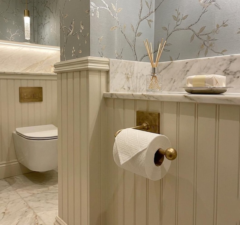 May include: A bathroom interior featuring a wall-mounted white toilet with a brass flush plate, a white toilet paper roll on a brass holder, and a glass reed diffuser on a marble shelf against cream-colored beadboard paneling and floral wallpaper.