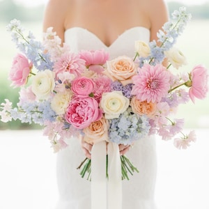 Puede incluir: Un vibrante ramo de novia con una mezcla de flores rosas, melocotón y blancas. El arreglo incluye rosas, dalias y otras flores, acentuado con flores azules y moradas. Cintas blancas caen en cascada desde la base del ramo.