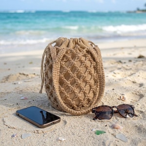 May include: A tan drawstring bag featuring a crocheted net overlay with a solid fabric interior. The bag is positioned on sand next to a black smartphone and dark-tinted sunglasses with brown frames, showcasing a textured, woven design.