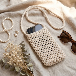 May include: A cream-coloured crochet phone pouch with a long matching shoulder strap, featuring an open-weave lattice pattern. The pouch contains a smartphone with a dark screen. The design is simple, functional, and crafted from cord.