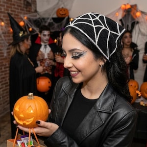 May include: A black triangular headscarf featuring a white crocheted spiderweb pattern. The headscarf is worn tied over dark hair, contrasting with the black fabric. The design consists of interconnected white lines forming a web across the black base.