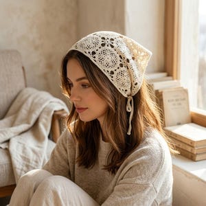 May include: Cream-colored, crocheted headscarf with a floral pattern. The head covering is tied under the chin with long, delicate strings. The scarf is made of a soft, open-weave material.