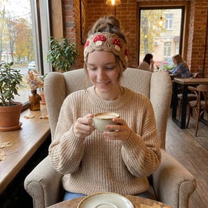 May include: A woman wearing a beige knit sweater and a crocheted headband with red mushroom designs. She is holding a cup of coffee in a cafe setting. The background includes a window, brick wall, and plants.