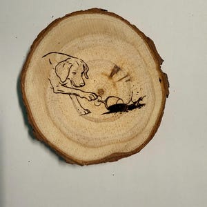 Może przedstawiać: Okrągły drewniany podkładka z ilustracją psa i rozlanego wina. Pies jest przedstawiony sięgając po kieliszek wina, a rozlane wino tworzy ciemną plamę. Podkładka ma naturalne usłojenie drewna.