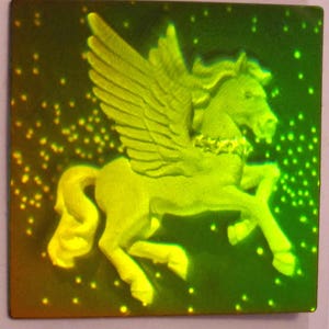 Puede incluir: Una pegatina hologr&aacute;fica verde y amarilla con un caballo alado con un collar dorado. El caballo est&aacute; representado en una postura erguida con las alas extendidas.