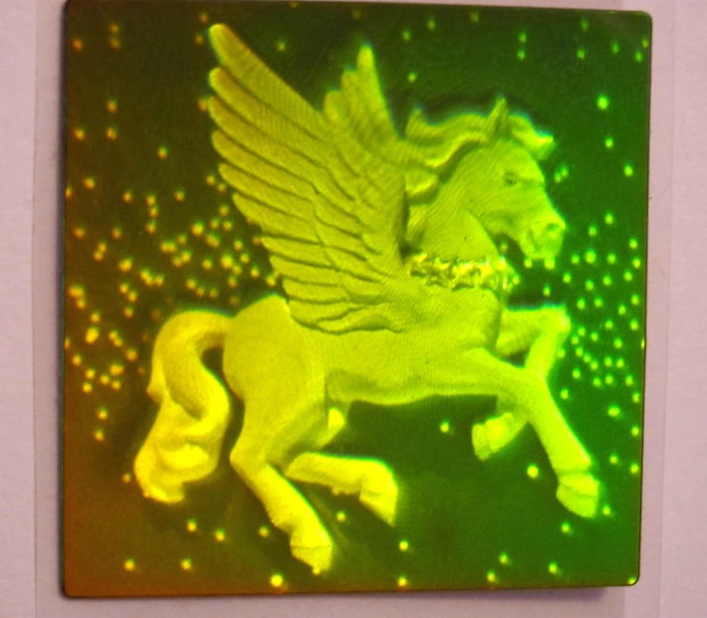 Puede incluir: Una pegatina hologr&aacute;fica verde y amarilla con un caballo alado con un collar dorado. El caballo est&aacute; representado en una postura erguida con las alas extendidas.