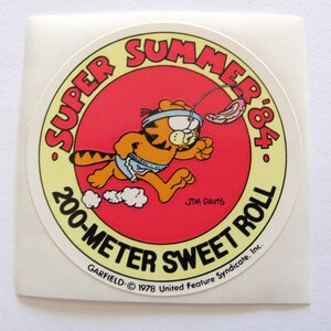 May include: A round sticker featuring Garfield the cat running with a red, white, and blue ribbon around his head. The sticker is yellow with red text that says "Super Summer '84 200-Meter Sweet Roll" and "Garfield &copy; 1978 United Feature Syndicate, Inc."