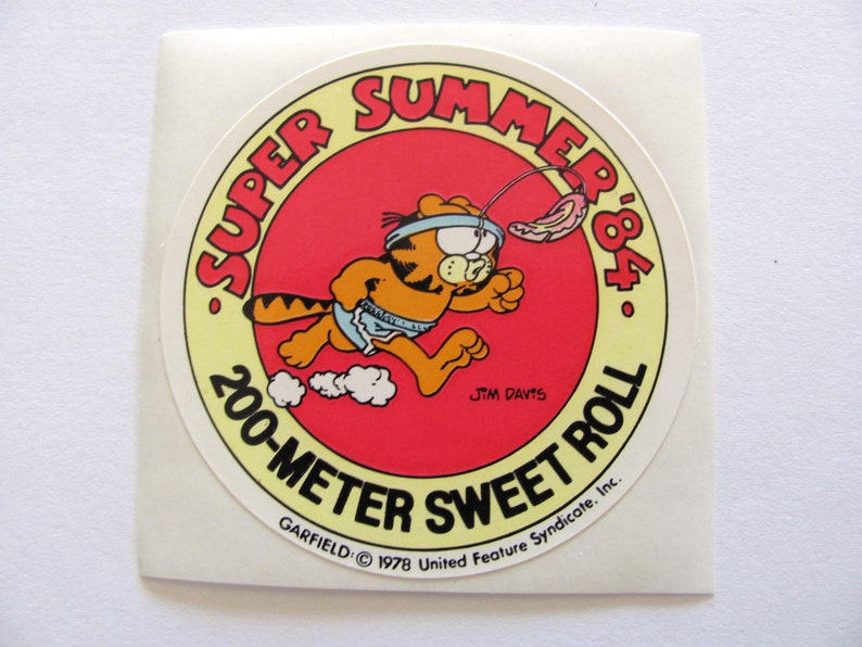May include: A round sticker featuring Garfield the cat running with a red, white, and blue ribbon around his head. The sticker is yellow with red text that says "Super Summer '84 200-Meter Sweet Roll" and "Garfield &copy; 1978 United Feature Syndicate, Inc."