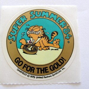 May include: A round, gold sticker with a cartoon cat, Garfield, wearing a number one ribbon. The sticker says "Super Summer '84" and "Go for the Gold!" with the Garfield copyright information at the bottom.