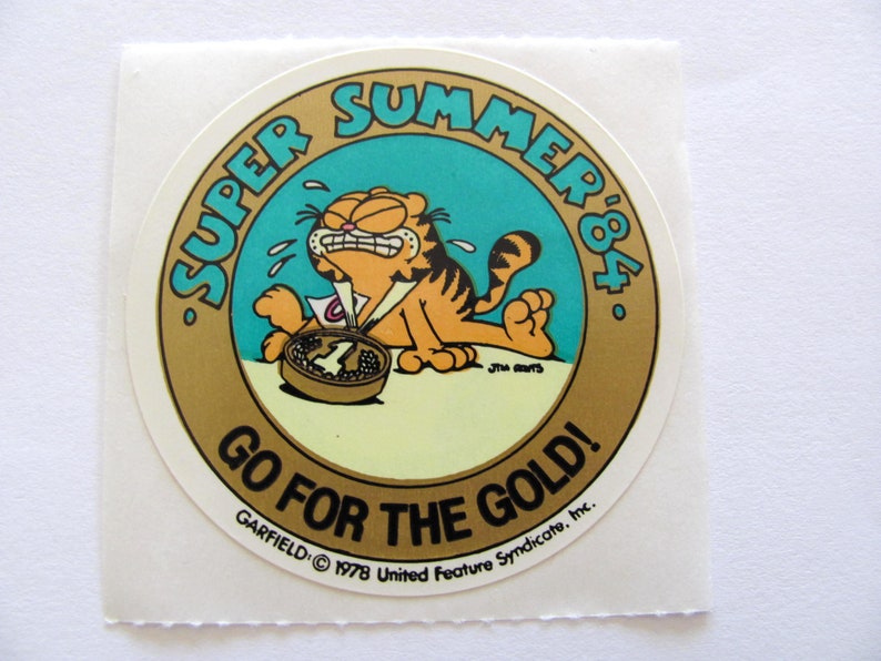 May include: A round, gold sticker with a cartoon cat, Garfield, wearing a number one ribbon. The sticker says "Super Summer '84" and "Go for the Gold!" with the Garfield copyright information at the bottom.