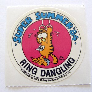 May include: A round sticker featuring Garfield the cat hanging from a set of gymnastic rings. The sticker is pink with a white border and the text "Super Summer '84" in blue. The text "Ring Dangling" is in blue and white and the text "Garfield &copy; 1978 United Feature Syndicate, Inc." is in black.