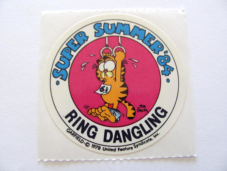 May include: A round sticker featuring Garfield the cat hanging from a set of gymnastic rings. The sticker is pink with a white border and the text "Super Summer '84" in blue. The text "Ring Dangling" is in blue and white and the text "Garfield &copy; 1978 United Feature Syndicate, Inc." is in black.