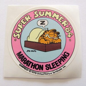 May include: A round sticker featuring Garfield the cat sleeping in bed. The sticker is pink with white text that reads "SUPER SUMMER '84. GARFIELD: &copy; 1978 United Feature Syndicate. Inc. MARATHON SLEEPING".