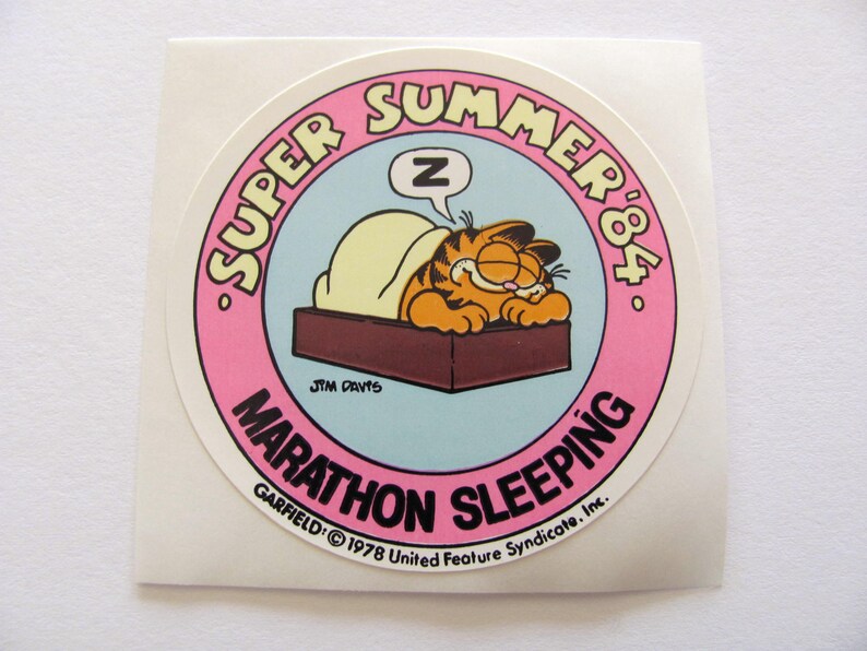 May include: A round sticker featuring Garfield the cat sleeping in bed. The sticker is pink with white text that reads "SUPER SUMMER '84. GARFIELD: &copy; 1978 United Feature Syndicate. Inc. MARATHON SLEEPING".