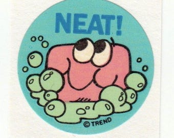 Könnte beinhalten: Ein runder Aufkleber mit hellblauem Hintergrund, der das Wort "NEAT!" in blauer Schrift zeigt. Eine rosa Cartoonfigur mit großen Augen ist von grünen Blasen umgeben. Der Aufkleber hat das "TREND"-Logo.